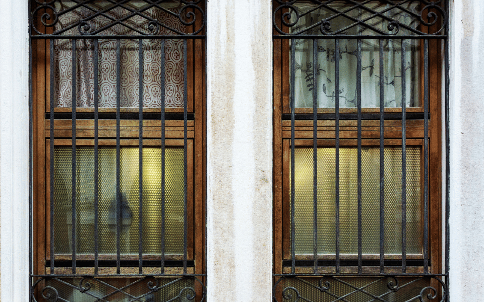 Ventana con rejas de seguridad verticales y horizontales de metal decorativo, instalada en una pared blanca. La ventana tiene un marco de madera y está dividida en dos secciones, con cortinas decorativas detrás.