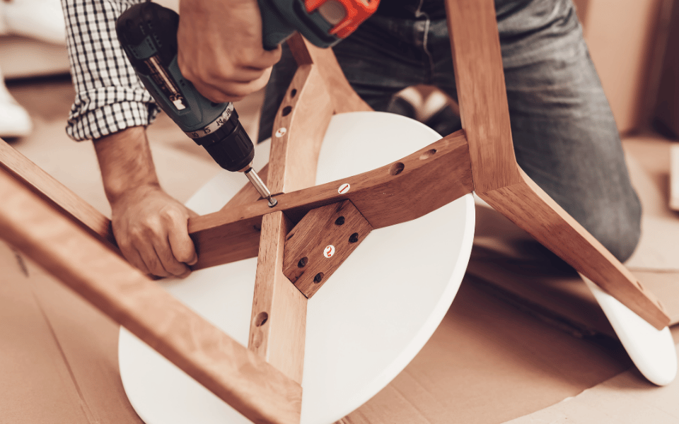 Profesional realizando una reparación de carpintería en una silla de madera, utilizando un taladro para asegurar un ensamblaje sólido y preciso.