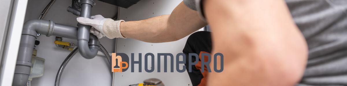 Plomero realizando el desazolve de un drenaje bajo un fregadero, utilizando guantes y herramientas profesionales. Logo de HomePro visible.